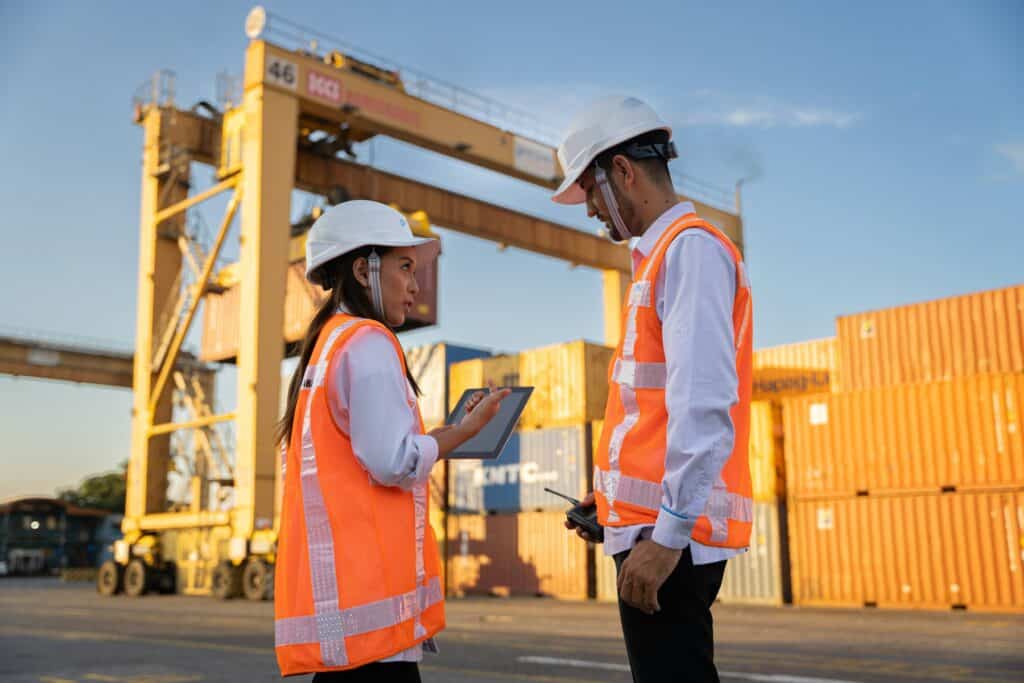 Two logistics workers in hard hats reviewing plans near shipping containers